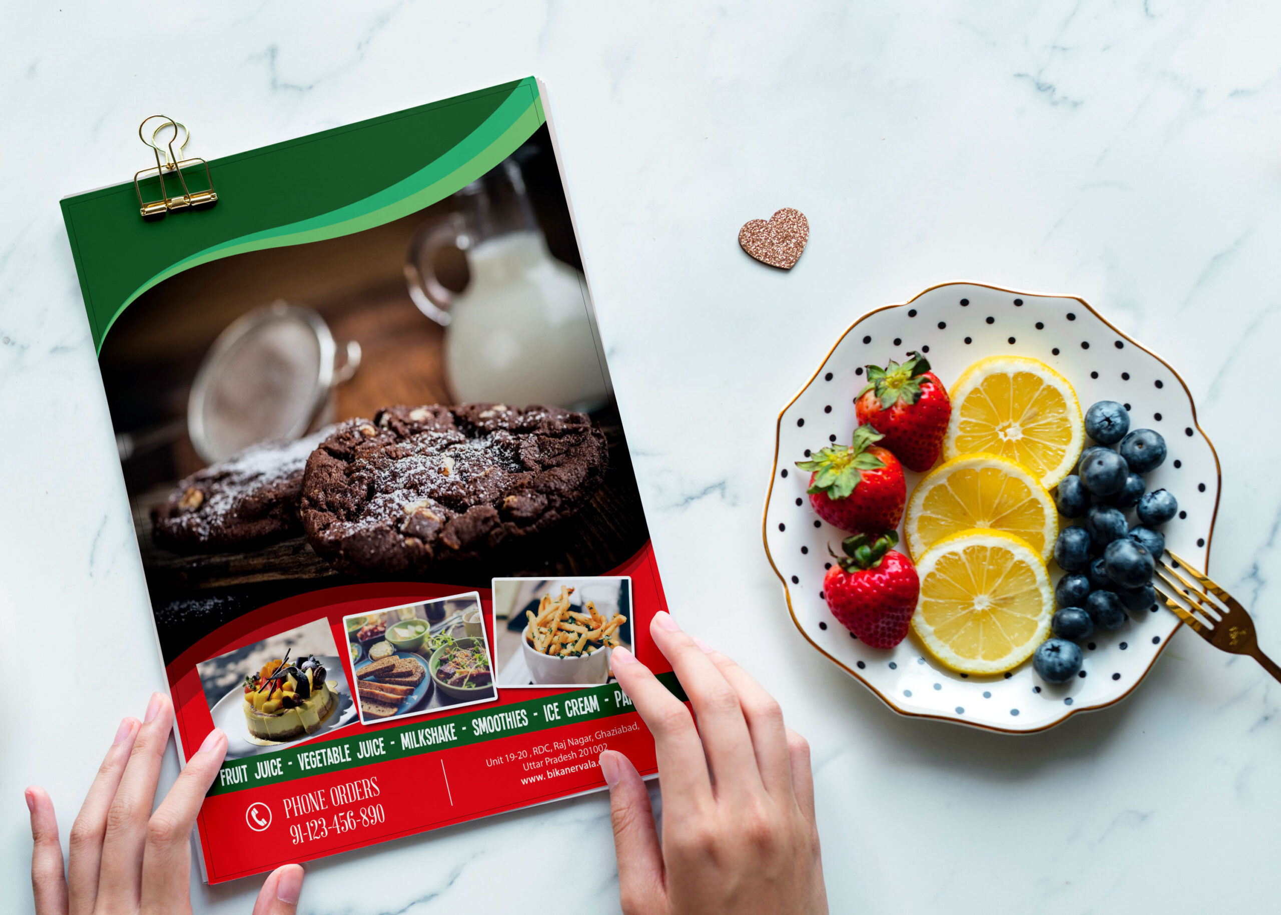 A hands holds a colorful bakery menu beside a plate of fresh strawberries, blueberries, lemon slices, and a gold fork.