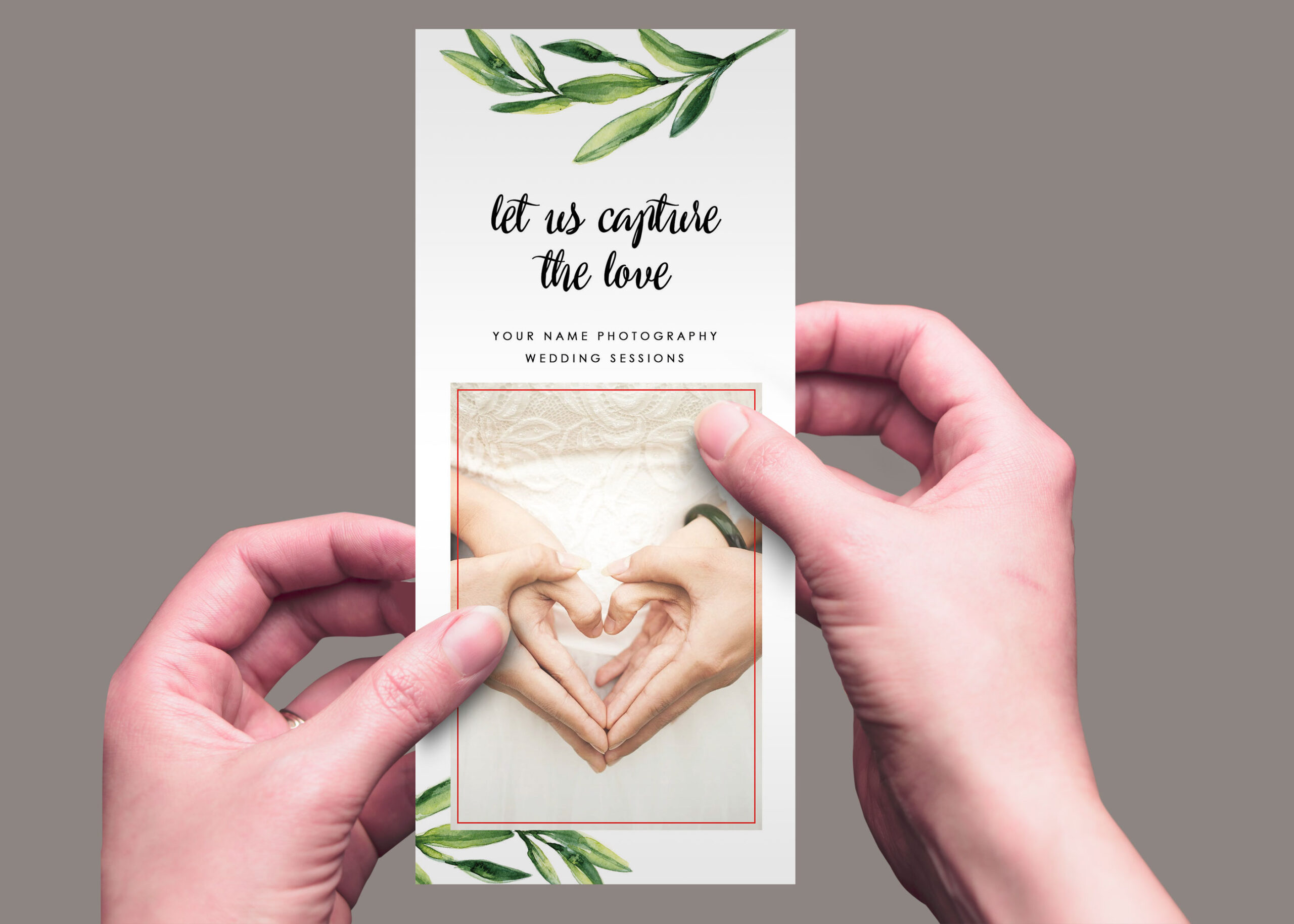 A person holds a wedding photography rack card featuring hands forming a heart, with green leaves in the design.